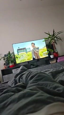 A low-angle, indoor shot shows a television screen with a man speaking on the phone. The television is mounted on a cabinet, behind which are plants and other objects. In the foreground, a grey blanket is spread across a bed. The room is dimly lit, with the television screen providing the primary light source.