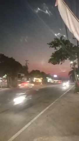 The video captures a twilight scene along a paved road. The sky displays a gradient of dark blue to pinkish-orange hues, indicating either dawn or dusk. Along the right side of the road, a white flag or banner is visible, attached to a pole. Trees line the left side of the road, with some buildings and structures interspersed. The road itself is wet, suggesting recent rain or a humid environment. Vehicles are in motion, their headlights and taillights creating streaks of light due to the long exposure or camera movement, blurring their forms. These streaks of light are indicative of cars driving on the road. The overall atmosphere is tranquil, illuminated by the fading natural light and the artificial lights of the vehicles and structures.