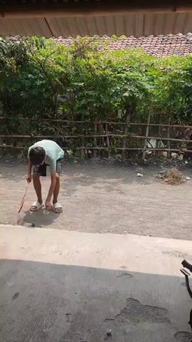 A young boy wearing a light blue t-shirt and black shorts is playing badminton. He is standing on a dirt path, with a badminton racket in his left hand and a shuttlecock in his right hand. He tosses the shuttlecock into the air and jumps to hit it with his racket. The background is a line of green bushes and a wooden fence. There is a roof overhead, casting a shadow on the ground.