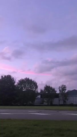 A time-lapse video shows a slow pan across a rural landscape at dusk. The sky transitions from a light purple to a deeper blue, with scattered clouds catching the last rays of sunlight, tinging them with pink. Trees and houses are visible in the mid-ground, along with streetlights and a road. A tall transmission tower is also present in the distance. The grass in the foreground is green, suggesting it is springtime or summer. There are no people or animals visible in the video.