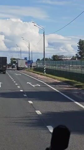 THE VIDEO SHOWS A VIEW FROM THE DRIVER'S SEAT OF A VEHICLE DRIVING DOWN A ROAD. A SERIES OF TRUCKS ARE VISIBLE, SOME PARKED ON THE SIDE OF THE ROAD AND SOME DRIVING. ON THE RIGHT SIDE OF THE ROAD, THERE IS A SOUND BARRIER FENCE. THERE ARE STREET LIGHTS AND UTILITY POLES ALONG THE ROAD. THE SKY IS BLUE WITH WHITE CLOUDS. ARROW MARKINGS ON THE ROAD INDICATE LANE CHANGES AND TURNS. A MICROPHONE IS VISIBLE AT THE BOTTOM OF THE FRAME, SUGGESTING THE VIDEO IS RECORDED FROM A VEHICLE WITH A DASHCAM OR SIMILAR DEVICE.