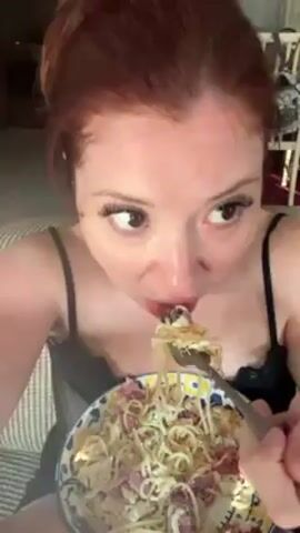 A pale-skinned woman with auburn hair piled on top of her head is holding a bowl of spaghetti with bits of what appears to be salami. She is looking down into the bowl, then looks upwards slightly, eating the spaghetti from a fork. She has on a black spaghetti-strapped shirt with black lace trim. She appears to be indoors, sitting on a striped sofa. Behind her is a wooden chair, a playpen, and a window. There is the sound of someone speaking in the background.