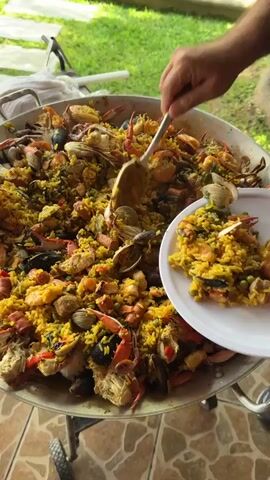 A person uses a large serving spoon to transfer paella from an extra-large paella pan onto a white plate. The paella contains seafood, including crab, clams, and shrimp, mixed with yellow-tinted rice and peas. The paella pan sits on a wheeled stand outdoors, with a green lawn visible in the background. The person's hand and part of their arm are visible, showing fair skin. The camera angle is slightly overhead, focusing on the pan and plate.