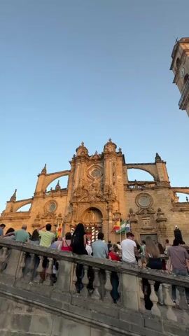A low-angle, vertical video shows a crowd of people sitting on a stone staircase, facing a large, ornate cathedral bathed in golden afternoon sunlight. The cathedral features intricate architectural details, arched windows, and a prominent central entrance. Flags are visible on the facade of the cathedral. The sky is clear blue. The people in the foreground are mostly seen from the back, dressed in casual clothing. Some are looking towards the cathedral, while others are engaged in conversation. The overall atmosphere appears relaxed and observational.