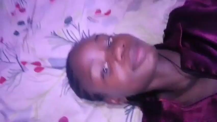 A person with dark skin and braided hair is lying on a bed with a patterned bedsheet. They are wearing a dark maroon or burgundy satin pajama top. They are looking at the camera. They put in earbuds. The video is recorded in a low-lit environment, giving the footage a purplish hue.