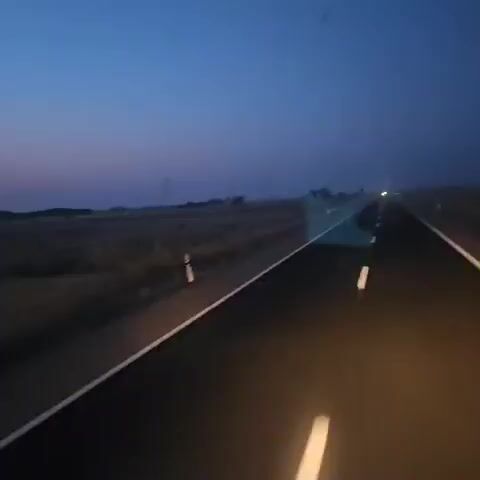 The video shows a view from a vehicle moving along a road during twilight. The sky transitions from a deep blue at the top to shades of purple and pink closer to the horizon. In the distance, there are fields of dry grass or crops and a few utility poles. The road has white dashed lines on the right side, indicating it's a lane divider. The interior of the vehicle is partially visible, with dark frames and what appears to be a side mirror reflecting the sky and a navigation device with a map displayed. The overall atmosphere is serene and suggests travel during the early morning or late evening.
