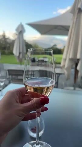 A person holds a wine glass with a small amount of rose wine in it. The hand has red nail polish. In the background are tables and chairs under large umbrellas on a patio. Beyond the patio is a green lawn with trees and mountains in the distance. The sky is blue.