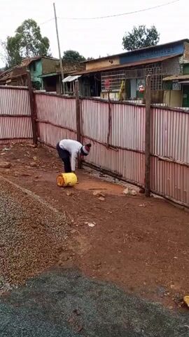 A person is working near a makeshift fence made of corrugated metal, propped up with wooden stakes. The ground is muddy. There's a yellow bucket lying on its side and it appears the person is stirring something in a puddle next to it.  To the left of the person is a pile of small stones.  In the background, there are buildings; one has a sign that says "DELY ZAGROVET" and another says "JASKOS HARDWARE". The sky is overcast.