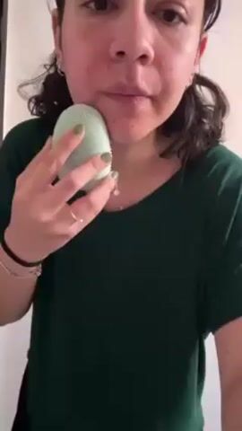A person in a green shirt is holding a green and white oval device against their face and moving it around, seemingly cleaning or exfoliating the skin. The person has dark hair and is wearing jewelry including a necklace, rings, and bracelets. The background appears to be a plain white wall.