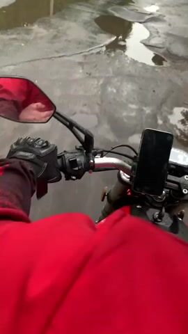 The video shows a first-person perspective of someone riding a motorcycle on a wet, paved road. The rider is wearing a red jacket and black gloves. The handlebars and a phone mounted on the motorcycle are clearly visible. The road is covered in puddles, indicating it is raining or has recently rained. Trees line the sides of the road, and buildings and parked cars are visible in the background. The lighting suggests it is daytime, possibly overcast due to the weather. The rider's reflection is visible in the motorcycle's mirror, showing a glimpse of their face. The overall atmosphere is one of a commute or journey in challenging weather conditions. There is no visible text in the video.