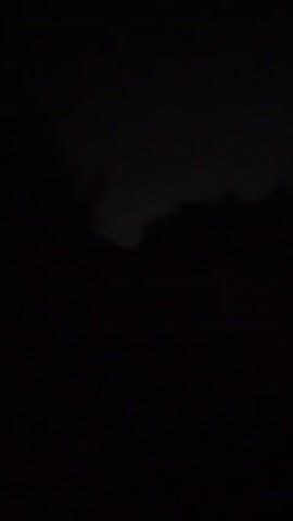 The video is dark and shaky, suggesting it was filmed at night. There are glimpses of trees or foliage silhouetted against a faintly illuminated sky. Flashes of light, likely lightning, intermittently illuminate the scene, revealing faint cloud formations and the outlines of trees. The flashes are brief and the darkness quickly returns.