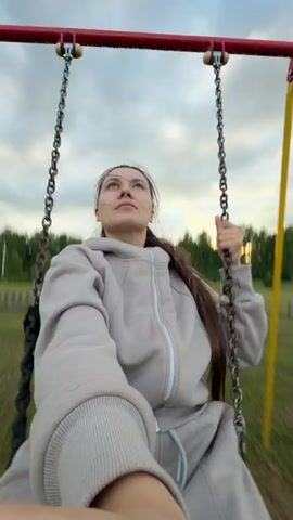A woman is swinging on a playground swing set outdoors. She is wearing a light gray hooded tracksuit and has long brown hair pulled back in a ponytail. The swing set has a red metal frame with yellow legs and chains. The background shows a grassy field with a wooden fence and trees in the distance, under a cloudy sky. The woman is holding onto the chains of the swing with both hands and looking towards the camera, with a neutral expression initially, then smiling as she swings. The camera appears to be held by the woman, suggesting a selfie-style shot. The lighting suggests it is either early morning or late afternoon.