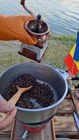 A person wearing an orange oven mitt is holding a vintage manual coffee grinder over a metal pot filled with roasted coffee beans. The grinder is being tilted to pour the beans into the pot. A wooden spoon is in the pot, stirring the beans. The scene appears to be outdoors, with a body of water in the background. A Romanian flag is visible on the right side of the frame. The pot is on a portable gas stove.