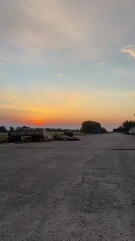 A vertical, full shot captures an outdoor scene at dusk. The sky displays a gradient of colors, transitioning from pale blue at the top to a vibrant orange and yellow near the horizon, where the sun is setting behind a silhouetted tree line. In the foreground, a vast expanse of gravel covers the ground. A pile of assorted objects, including what appear to be stacked pallets and possibly scrap metal, are situated on the left side of the frame. Further back, a dark-colored truck bed is visible. On the right side, a blue tarp is draped over an unseen object, and a lone tree stands against the horizon. The overall impression is one of rural quietude as day turns to night.