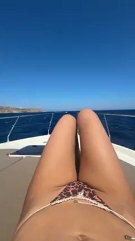 Here is a description of the video:

The video shows a person relaxing on the front of a moving boat. The individual is lying down with their legs towards the camera, wearing a bikini. The clear blue sky and the ocean's surface are visible in the background. As the video progresses, the boat moves, and the view shifts to show the distant shoreline and the open sea. The sun is shining brightly, adding to the overall bright and sunny atmosphere. The boat moves swiftly through the water, causing waves and splashes.