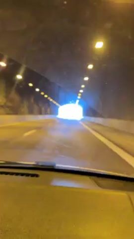 The video shows a car driving through a tunnel. The tunnel has lights overhead and appears to be made of concrete. Other vehicles are visible in the distance, also traveling through the tunnel. The end of the tunnel is brightly lit. The footage is taken from inside the car, looking out through the windshield.