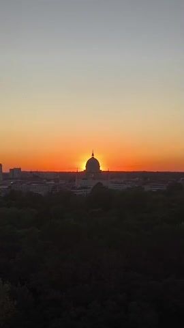 This video displays a sunset over a cityscape. The sun is setting behind a domed building, casting an orange glow across the sky. In the foreground, there is a dark expanse of trees. Buildings of various heights are visible in the distance to the left of the domed structure. The sky transitions from a light grey at the top to a vibrant orange near the horizon. The video appears to be a static shot, with no discernible movement within the frame.