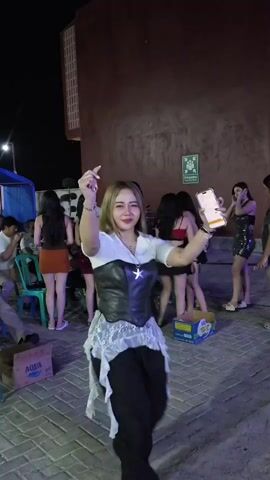 A young woman with blonde hair, wearing a white shirt, black corset with a star detail, and black pants, dances in front of a group of women standing in a line. She is holding a phone in her right hand and gesturing with her left hand. The background features a reddish-brown wall, a green sign, and brick pavement. Several bottles of water are visible in a box on the ground. The scene appears to be outdoors at night.