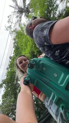 The video shows two people, a man and a woman, with luggage on a street that is lined with palm trees and lush green vegetation. The camera is held from the perspective of the person in the foreground, who is holding a green suitcase. The woman is in the foreground, wearing a red bikini and a pearl necklace, smiling at the camera and holding the suitcase. The man is behind her, wearing glasses and a black and gray patterned shirt, looking upwards and to the right. The man appears to be holding onto the suitcase or the woman, as they are positioned close together. The scene appears to be outdoors, possibly in a tropical or subtropical location, with a cloudy sky visible. The ground is a paved surface, and the foliage suggests a warm climate. The overall impression is that they are traveling, possibly to or from an airport or a vacation destination.