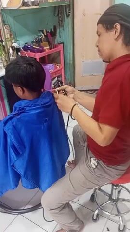 A person with dark hair wearing a red shirt and tan pants is sitting on a red stool and cutting the hair of a young person sitting in a chair. The young person is wearing a blue cape. The barber is holding a comb and clippers in their hands and is actively cutting the hair of the young person. The background appears to be a room with shelves holding various items, possibly a salon or barber shop.