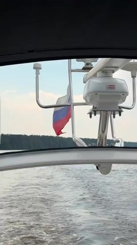 The video is shot from the deck of a moving boat, looking towards the stern. A Russian flag waves from a pole on the right side of the frame. In the background, a wooded shoreline is visible under a hazy sky. A large white satellite dome with "TRACVISION KVH" written on it is mounted on a metal pole towards the right side of the frame. The water is choppy, creating a wake behind the boat.