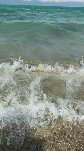 The video shows a close-up, low-angle view of gentle waves breaking on a rocky shore. The water is a murky green-blue, and the seabed is visible through the shallow water, littered with pebbles and stones. The waves create white foam as they wash ashore, and some debris floats on the surface. In the distance, the sea stretches out to a hazy horizon under a bright, cloudy sky. The scene is peaceful and natural, capturing the movement and sound of the ocean.