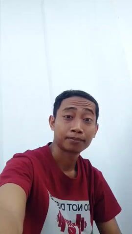 A man with short black hair and brown skin is wearing a red t-shirt and looking at the camera. He is sitting against a white wall. The t-shirt has a white graphic and text on it. The lighting is bright.