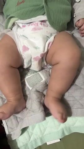 The video shows a close-up of a baby's legs and diaper. The baby is lying on a quilted surface. The baby's legs are chubby and the baby is wearing a diaper with a cloud pattern. The baby's feet are visible and moving slightly. The baby is wearing a green shirt or onesie. The background is a softly lit room.