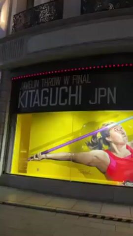 Here is a description of the video:
The video shows an artistic window display for the Javelin Throw Women's Final, featuring Kitaguchi from Japan. The display includes a mural-like image of Kitaguchi in a red athletic top, in the motion of throwing a purple javelin. The background is yellow. Above the image is text that reads "JAVELIN THROW W FINAL KITAGUCHI JPN." To the right, the number "67.73" is displayed in a stylized font. The display is in a window of a building with ornate architectural details above. The sidewalk is visible below the window. The video is taken at night.