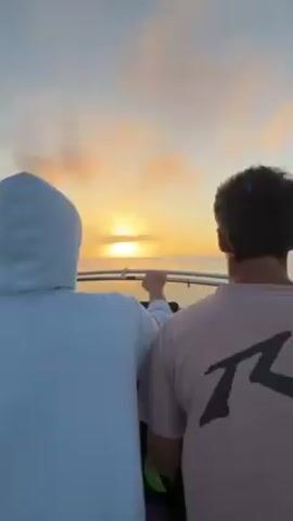 The video opens with a static shot of three men standing on a boat, looking towards the horizon at the setting sun. One man is wearing a white hooded sweatshirt and points towards the sun. The next shot pans slightly to the side, revealing a fishing rod and the ocean. The camera then shifts to the back of the boat, showing the wake created as the boat moves quickly through the water. A Honda 135 engine is visible. The sky is a pale blue with some light clouds.