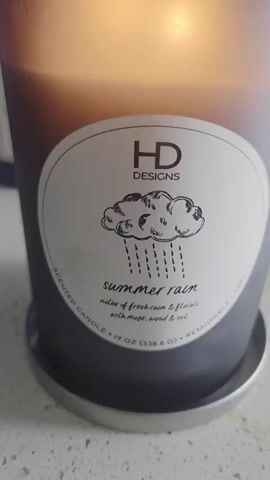 A close-up video shows a lit HD Designs Summer Rain scented candle. The candle is in a frosted gray jar with a silver lid. The label on the jar has a drawing of a cloud with rain falling, the words "HD DESIGNS", "summer rain", and "notes of fresh rain & florals with sage, wood & soil." The video shows the flickering flames of the candle and the candle wax melting. The candle is sitting on a white speckled countertop with a silver coaster.