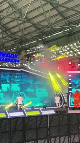 This is a video clip of a stage performance in what appears to be an indoor venue with a large, industrial-style roof structure. The stage is set up with large screens displaying images of cars and what looks like code or digital graphics. Bright stage lighting, including red and yellow spotlights, illuminates the scene, and there is some smoke or haze.

The subjects on stage include several people dressed in bright orange jumpsuits and helmets, and two humanoid robots in white outfits. They appear to be part of a coordinated performance, with the people in orange moving in unison. One robot is positioned on the left side of the stage, and another is towards the center.

A large banner with blue lettering is visible at the top of the stage, with the text "DDK" clearly legible. The background screens show stylized images of cars, suggesting a theme related to automotive or technology.

The overall impression is of a modern, high-tech performance or product launch event. The lighting and presentation indicate a lively and energetic atmosphere. There are no clear indicators of the time of day or weather conditions, as the event is indoors. The location is specified as Dadukou, China.