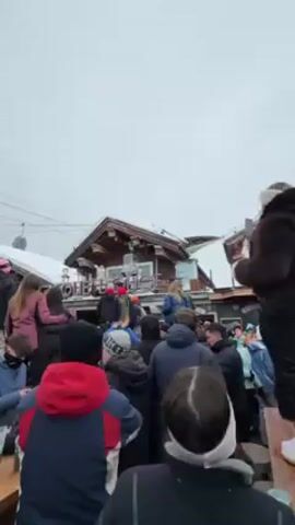 The video shows a crowd of people at an outdoor apres-ski venue. They are gathered around a wooden building with a sign that says "Die Drehleier". People are drinking and socializing. The camera is held by a woman wearing glittery makeup, who turns the camera on herself and a man in a black and white checkered jacket. They both pose for the camera, waving. The woman and man kiss. The scene is snowy and the sky is overcast. A few people are on top of the building in the background. There are beer glasses on a table in the foreground.