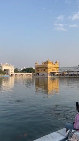 A vertical shot shows a wide view of the Golden Temple reflected in a large body of water. The golden structure stands prominently in the center, with a pathway leading to it. People are visible walking on the pathway and near the water's edge. In the foreground, a person wearing a hijab and pink clothing is sitting on the edge of the water, facing away from the camera and towards the temple. The sky above is clear with a few wispy clouds. The water's surface has ripples, and some faint circles indicate movement below the surface, possibly from fish or disturbances.