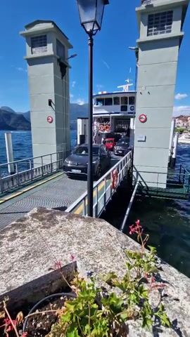A dark-colored SUV is positioned on a ferry ramp, facing forward towards the ferry boat. The ramp is connected to the ferry, which is visible in the background. Two gray towers with square tops stand on either side of the ferry ramp, flanking the entrance to the ferry. A black lamppost is situated between the towers. The ferry itself appears to be a passenger and car ferry, with a white upper deck and visible windows. The water surrounding the ferry ramp is a deep blue, and the sky above is clear and bright blue. In the foreground, a stone ledge with a potted plant containing green leaves and pink flowers is visible, partially obscuring the view of the ramp and ferry. The overall scene suggests a ferry terminal on a sunny day.