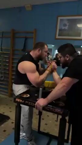 Two men are arm wrestling on a table with “ARM FIGHT FACTORY” printed on it. They are wearing black shirts and are secured together by yellow wrist straps. The man on the left has short hair and a tattoo on his left forearm. The man on the right has a beard. They are both staring intently at each other. There are other people in the background, and a ladder leaning against the wall. There is also a framed picture hanging on the wall.