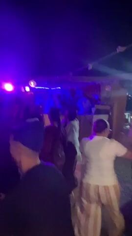 The video shows a nighttime outdoor party with a crowd of people dancing under blue and red lights. A DJ booth is visible in the background with a DJ performing. Some people are blurred due to movement. The atmosphere appears lively and celebratory.