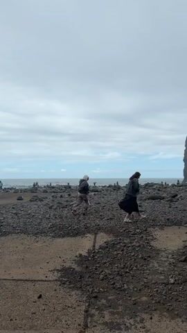 Two people are walking on a rocky beach towards the ocean. The beach is covered in small dark rocks and some sand. In the background, the ocean is visible under a cloudy sky. In the far distance, there are small rock towers. A large rock formation is visible on the right side of the frame. One person is wearing light-colored pants and a dark jacket, and has a camera. The other person is wearing a dark skirt or dress and a hooded jacket.