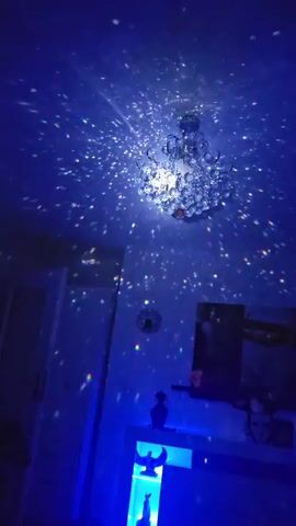 The video shows a dimly lit room with blue lighting. A chandelier hangs from the ceiling, casting scattered light patterns on the walls and ceiling. There is a wall clock and a shelf with decorative items, including a statue with wings and a crescent moon. The lighting creates a sparkling, starry effect.