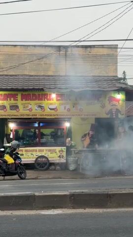The video shows a street-side food stall with a large yellow sign displaying "SATE MADURA CAK YUSUF" in red and yellow letters. The stall appears to be a simple cart with a covered roof. Smoke is rising from a grill area where a person is cooking. Several people are visible around the stall, including a person on a motorcycle to the left and individuals standing or sitting near the cooking area. Vehicles, including a turquoise car and other blurred motorcycles, are passing by on the road in front of the stall, indicating a busy street environment. The background shows a building with a yellow facade and a gray, overcast sky. The overall scene depicts a bustling urban setting with a focus on a local food vendor.