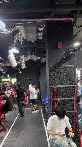 The video captures a bustling scene inside a gym. Several people are working out on red exercise machines, while others walk around the room. The gym has a dark ceiling with exposed ductwork and round fluorescent lights. A variety of gym equipment is visible throughout the space, including weight machines and free weights. One person is using a barbell. There are signs with Chinese characters on the walls.