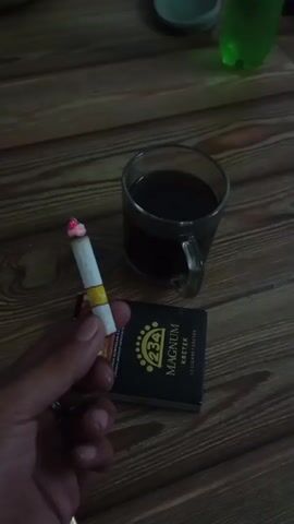 A person's hand holds a lit cigarette, positioned above a pack of Magnum Kretek cigarettes and a glass of dark liquid, likely coffee. The scene takes place on a wooden surface.