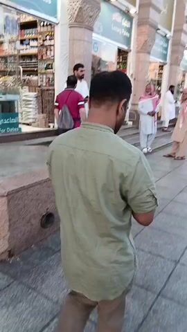The video shows a street scene in Saudi Arabia. Several men wearing traditional Arab clothing are standing outside of shops labeled "Luxury Palms" and "Good Palm for Dates." One man is wearing a light green shirt and khaki pants and walks in front of the camera. The shops are selling dates. The architecture is classical. The street is paved with light gray stone.
