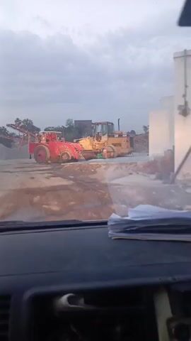 From the perspective of inside a car, the video shows two construction vehicles on a dirt lot. The vehicle closest to the camera is a yellow "CAT 928F" front-end loader. Beside it is a red, similarly shaped vehicle. The sky is overcast and pale. There's a pile of papers on the car's dashboard.