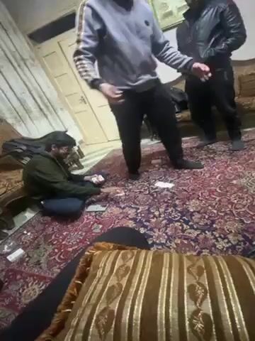 The video shows a living room with three men and one person filming. One man is seated on the floor with cards in his hand, while another is standing and moving around. The third man stands with his hands in his pockets, facing the other two. The room has a patterned rug and several pieces of furniture, including sofas and a small table. A window is visible in the background, and a guitar case rests against one of the sofas.