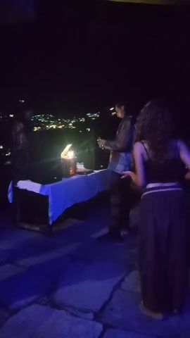 A video shows a nighttime outdoor gathering. In the foreground, a person with long curly hair is wearing a dark sleeveless top and loose-fitting pants. They are standing near a table covered with a white cloth, on which sits a lamp and some objects. Behind them, another person, appearing to be a man with dark hair, is wearing a patterned shirt and dark pants. He is holding a glass. The background reveals a distant town or city illuminated by lights, suggesting the gathering is situated on a vantage point overlooking the area. The ground appears to be paved with large stones. The scene is illuminated by artificial lighting, casting blue and yellow hues.