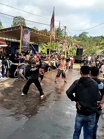 The video shows an outdoor cultural performance. In the foreground, several men are engaged in what appears to be a martial arts or dance demonstration. One performer wears traditional attire with a headdress. In the background, a crowd watches from the side of a wet, paved area. A stage with speakers and a banner is set up, suggesting a performance venue. Buildings, including one with a sign that reads "MASJID AL HIDAYAH", are visible. The environment is rural or semi-rural, with trees and utility poles in the distance. The overall scene depicts a lively community event.