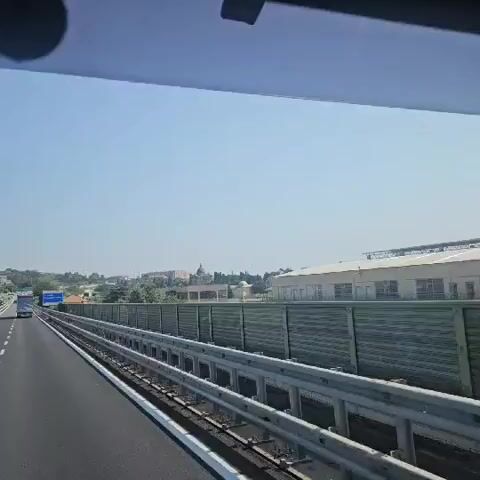 The video shows a view from inside a vehicle traveling on a highway. The camera pans slightly, capturing the road ahead and to the right. A guardrail runs along the side of the highway, and beyond that, there is a sound barrier. In the distance, there are buildings, trees, and a church with a dome. A blue sign is visible, indicating "citta SAVONA Aurelia del Colle di Cadibona 600 m". The sky is clear and blue, suggesting a sunny day.
