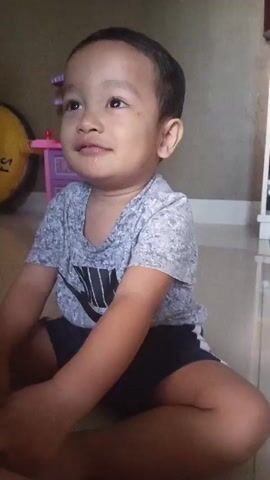 A young child sits on the floor in front of a pink toy kitchen set and a white shelving unit. The child is wearing a gray t-shirt with a black Nike logo and blue shorts. The child looks directly at the camera, and in the second half of the video, the child opens their mouth and speaks. There is a slight amount of saliva visible on the child's chin.