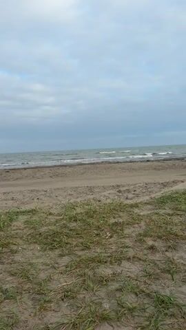 The video shows a beach scene. The sky is cloudy, and the sea is choppy. There is sand and sparse vegetation in the foreground. Two people are walking along the beach, and a blue car is parked nearby. The people appear to be interacting with each other.